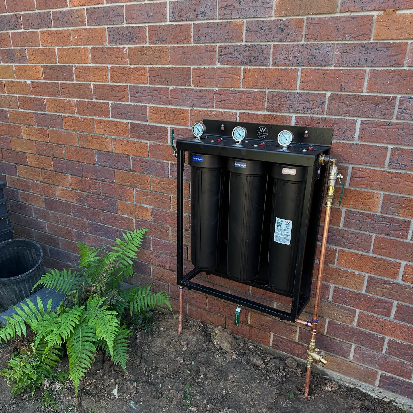 Three-canister water filtration system mounted on a brick wall with pressure gauges